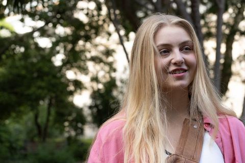 Young woman smiling in park wearing pink cardigan and brown leather crossbody strap, natural light