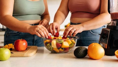 Preparing colorful fresh fruit salad in glass bowl by two women in bright modern kitchen