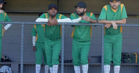 Diverse baseball team leaning on dugout railing wearing green yellow uniforms focused