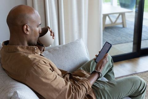 Relaxed african american man sipping coffee and scrolling smartphone on cozy sofa