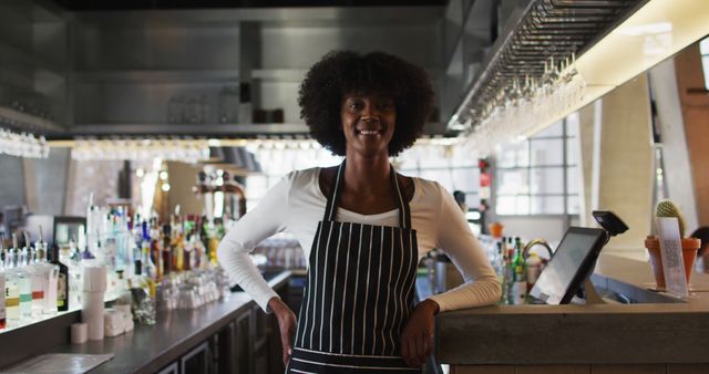 Smiling African American bartender standing confidently in a modern bar with hands on hips. Great for themes related to hospitality industry, bartender profiles, modern bar settings, customer service, and small business ownership.