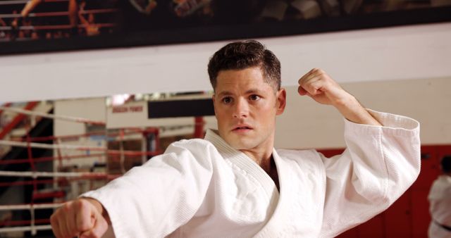 Young male martial artist wearing traditional white karate uniform, practicing punching techniques in gym. He is focused and determined, suggesting dedication to training and fitness. This image can be used to illustrate concepts of self-discipline, physical fitness, martial arts training, and self-defense methods.
