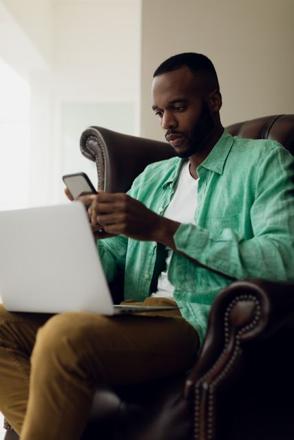 Side view of an African-American sitting on a leather chair inside a room while using a smartphone and a laptop on his lap