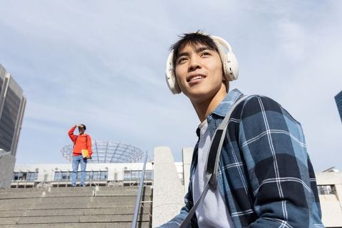 Young asian man listening to music on headphones sitting on urban steps with backpack