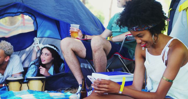 Group of friends enjoying time at campsite. Woman using smartphone, while others sit on mat and drum near tent. Ideal for content about outdoor activities, socializing, technology, travel, and summer adventures.