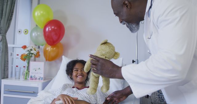 Diverse male doctor playing teddy bear with girl in hospital bed wearing fingertip pulse oximeter. medicine, health and healthcare services.