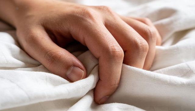 Hand gripping white bedsheet, intimate closeup of fingernails and fabric texture