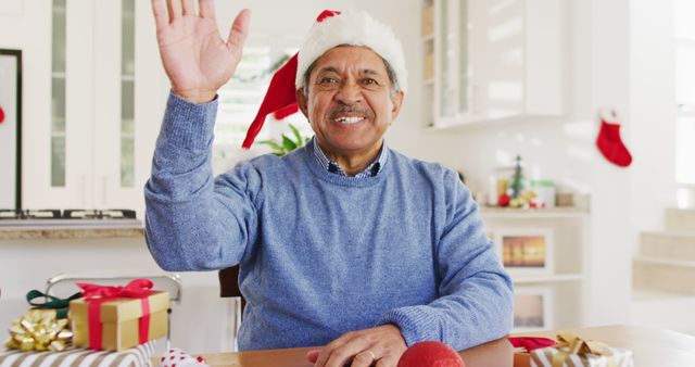 An elderly man, wearing a Santa hat and a blue sweater, waves while surrounded by wrapped gifts and holiday decorations in a bright, modern home. Perfect for ads, greeting cards, and social media posts celebrating the holiday season, family gatherings, and festive spirit.