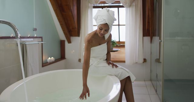 Woman is sitting on the edge of a bathtub, touching water in a bathroom. Wrapped in a towel with another towel on her head, she appears relaxed. Suggests peaceful quiet moments spent in personal leisure. Perfect for beauty and wellness promotions, spa advertisements, selfcare blog posts, or bathroom decor inspirations.