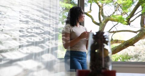 Curly-haired woman holding mug gazing out large window with sheer curtain, sunlit tree view