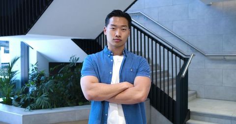 Asian man standing with arms crossed in modern stairwell projecting professional confidence