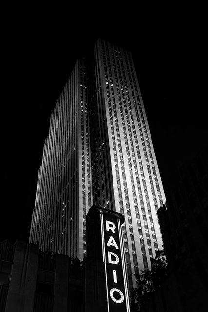 Striking view of a towering Midtown Manhattan skyscraper with a prominent 'RADIO' neon sign illuminated at night. Black and white tones highlight the timeless architecture and urban elegance. Perfect for concepts involving history, iconic landmarks, city life, and architectural grandeur.