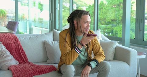 Man with dreadlocks speaking into smartphone while sitting on modern bright living room sofa