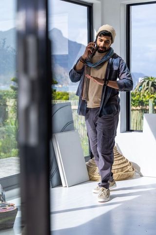 Indian man checking smartwatch while talking on phone in sunlit modern studio with mountain view