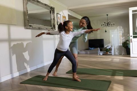 Asian mother guiding african american daughter practicing yoga in sunlit living room