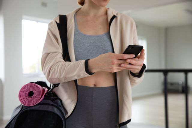Caucasian female ballet dancer in activewear holding a smartphone and fitness bag with a yoga mat in a bright ballet studio. Mirror and barre in the background. Ideal for use in fitness, dance, technology, and lifestyle contexts, such as promoting dance classes, fitness apps, or activewear brands.