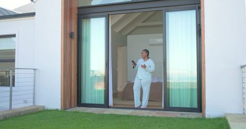 African american woman in pajamas standing at sliding door holding smartphone and mug