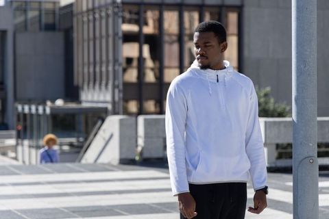 African American man standing on sunlit urban plaza wearing white hoodie contemplating city