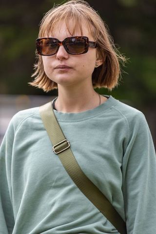 Young woman wearing tortoiseshell sunglasses and green sweatshirt with olive crossbody strap