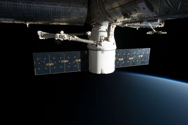 Image depicts a spacecraft docked to the International Space Station with solar panels extended, floating above the Earth's horizon in the vast blackness of outer space. Useful for articles and educational materials about space missions, space technology advancements, and the function of the ISS in scientific exploration.