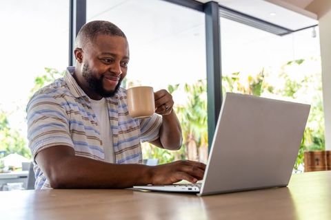 African man working remotely on laptop drinking coffee at modern bright home workspace