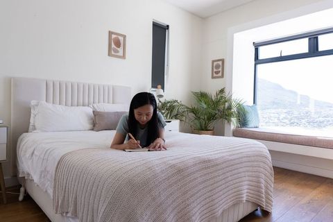 Mature african american woman writing in notebook on bed in bright minimalist bedroom