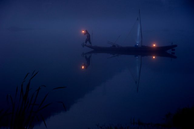 Fisherman standing on a traditional wooden boat holding a lit lantern during a foggy evening on a calm body of water. The scene creates a serene and mystical atmosphere with the soft light reflecting off the water's surface. Ideal for use in travel magazines, nature retreats promotions, themes of tranquility and solitude, and backgrounds for inspirational quotes.