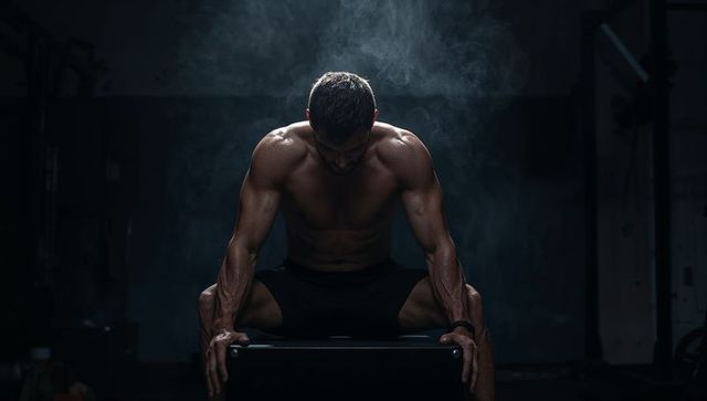 Sitting muscular athlete gripping workout box in dark gym spotlight, chalk rising