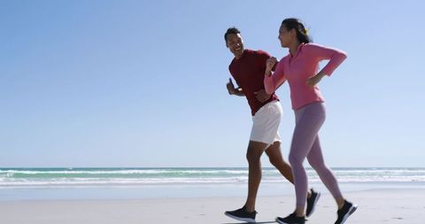 Diverse couple jogging on sunlit beach wearing fitness trackers and running shoes