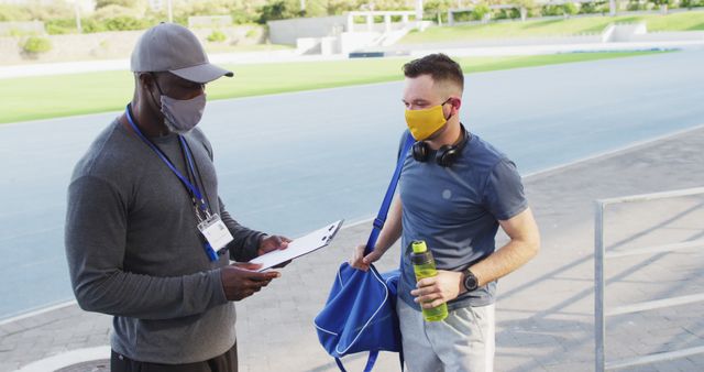 Athlete and coach wearing masks talking about training plan at outdoor track. Useful for fitness, sports, training programs, and health safety-themed content.