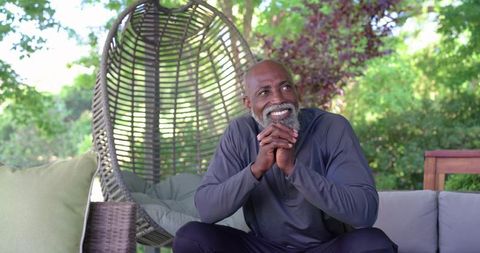 Senior african american man smiling on patio sofa by rattan chair, relaxed lifestyle