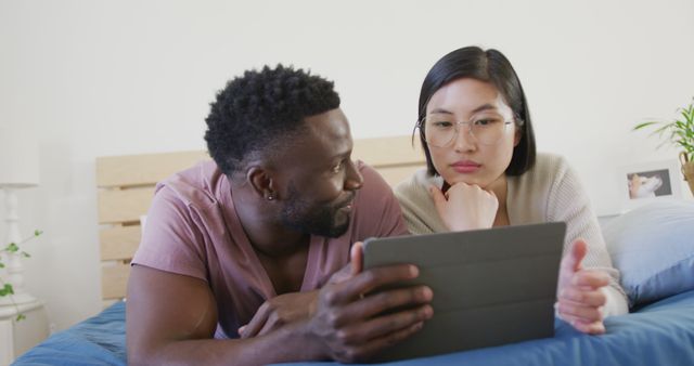 Diverse couple reclining on bed, using tablet device together. Can be used for lifestyle, communication, technology, or modern living contexts. Ideal for promoting tech accessories, apps, or web-based services involving couples or relationships.