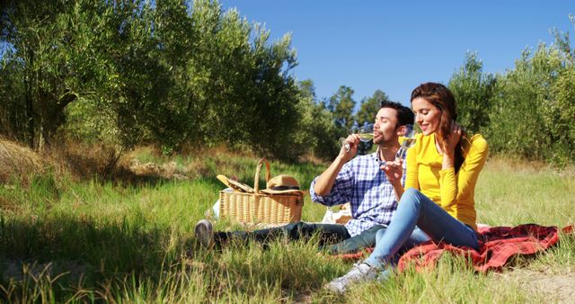 A Caucasian couple enjoys a sunny day outdoors, sitting on a red blanket and having a picnic, with copy space. They appear relaxed and happy, sharing a moment of leisure in a natural setting.