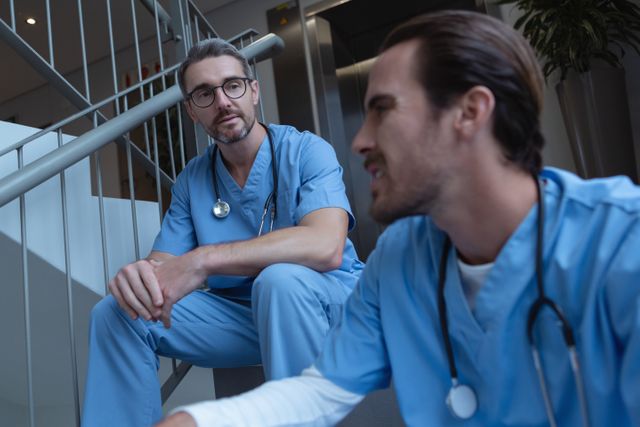 Two male medical professionals wearing blue scrubs and stethoscopes are having a discussion while sitting on a hospital staircase. Ideal for content related to healthcare, teamwork in medical settings, and hospital environments.