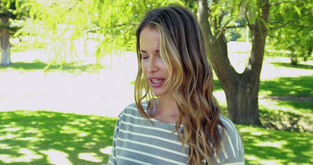 Woman enjoying a sunny day outdoors in a lush, green park. Her long hair and casual striped top convey a relaxed, happy atmosphere. Ideal for promoting outdoor activities, wellness, leisure, and fashion.