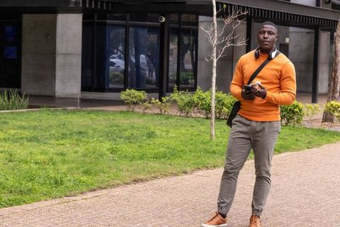 Young man walking campus with smartphone and snack wearing orange sweater and headphones