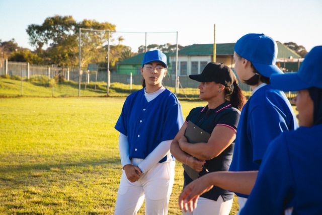 Female baseball players and their coach are discussing game tactics on a sunny day at the baseball field. The coach is holding a clipboard, and the players are wearing blue uniforms and caps. This image can be used for promoting women's sports, teamwork, athletic training programs, and diversity in sports.