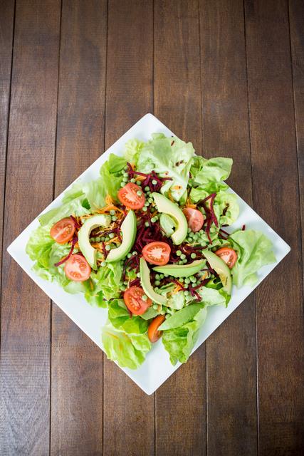 This image showcases a fresh vegetable salad with avocado slices, cherry tomatoes, and lettuce on a white plate placed on a wooden table. Ideal for use in health and wellness blogs, nutrition articles, restaurant menus, and food-related advertisements promoting healthy eating and vegetarian or vegan lifestyles.