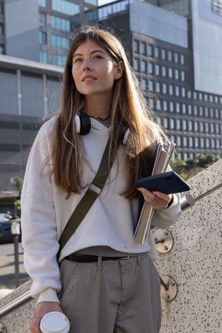 Young woman standing on urban steps holding notebooks, phone and coffee, city commuter