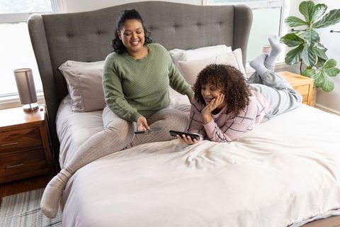 Diverse mother and teenage daughter relaxing on bed sharing smartphones and laughing together