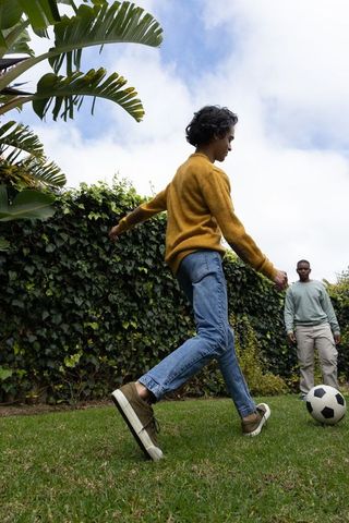 Male friends playing backyard soccer near lush hedge wearing mustard sweater and jeans