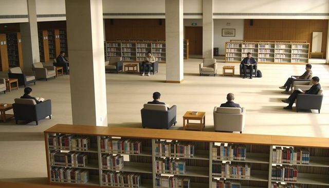 Spacious library reading room with adults sitting and reading in armchairs among shelves