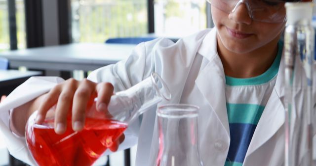 A boy wearing safety glasses and a lab coat is conducting a chemistry experiment, carefully pouring a red liquid into a glass flask. Ideal for educational content, children's science programs, STEM activity promotions, school presentations, and science-related websites.