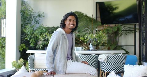 Young man in his 20s smiling and relaxing in bright sunroom wearing gray hoodie