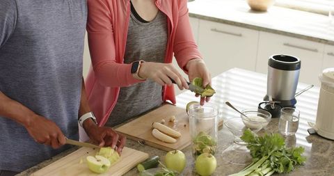 Couple preparing green smoothie in bright kitchen slicing apple and celery for blender
