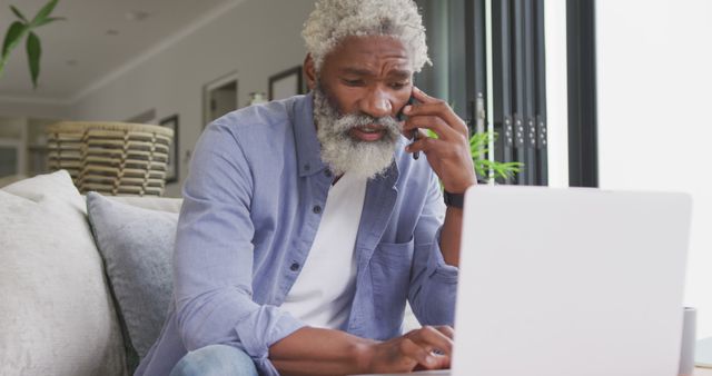 Elderly man with gray hair talking on phone while working on laptop in home office. He is wearing a casual blue shirt and jeans, multitasking between communication and computing. Ideal for illustrating remote work, senior technology use, and home office environments.