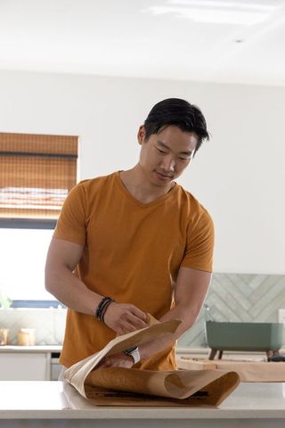 Asian man peeling parchment from freshly baked homemade bread on kitchen countertop