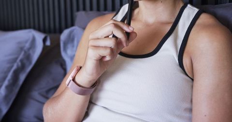 Thoughtful young woman reclining on bed holding pen wearing pink smartwatch and white tank