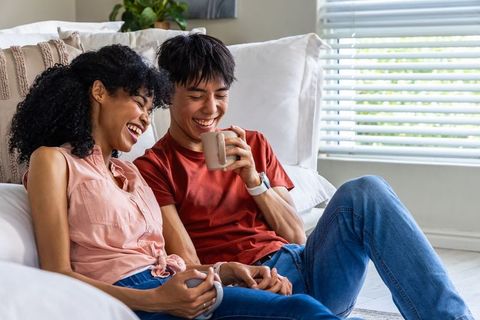 Laughing diverse couple sharing coffee and holding mugs on white sofa in cozy living room