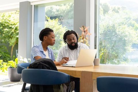 African mentor mentoring teenage woman studying on laptop at sunlit wooden table, greenery